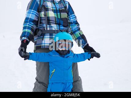 Un tout-petit garçon en combinaison bleu chaud, casque et lunettes apprend à skier, tient son père par les mains. Journée froide dans les montagnes enneigées, activités utiles avec Banque D'Images