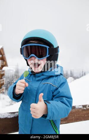joyeux petit garçon dans un casque, des lunettes et une combinaison bleue à la station de ski. Animations hivernales pour les enfants. Il mange un Lollipop, se montre comme avec on Banque D'Images