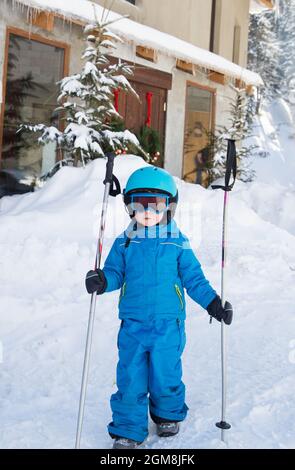 Un tout-petit garçon dans un casque, des lunettes, des skis et une combinaison bleue marche avec des bâtons de ski dans la station de ski. Préparation à la leçon de ski à l'école de ski. Hiver actif en Banque D'Images