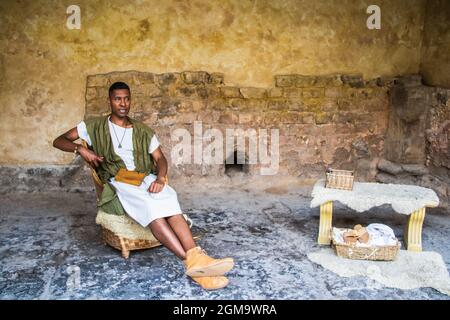 2019 07 25  Bath UK - réacteur noir dans les poses de costume comme érudit romain dans la salle de pierre avec livre en bois et table de travail à l'intérieur des bains Banque D'Images