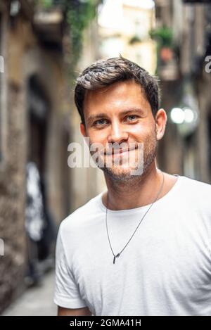 Contenu jeune homme barbu dans un t-shirt blanc décontracté souriant et regardant l'appareil-photo tout en se tenant sur la petite rue de la vieille ville en été Banque D'Images