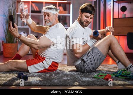 Homme d'âge moyen s'entraîne avec des haltères et souriant sur l'appareil photo. Retraité sportif utilisant un smartphone derrière. Formation domestique du père et du fils. Banque D'Images