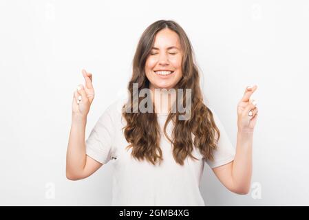 Une jeune femme pleine d'espoir croise ses doigts pour souhaiter quelque chose sur fond blanc. Banque D'Images
