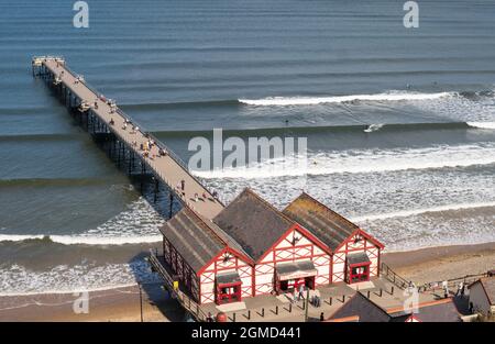 Personnes marchant le long de la jetée de Saltburn avec des surfeurs dans l'eau à côté, North Yorkshire, Angleterre, Royaume-Uni Banque D'Images