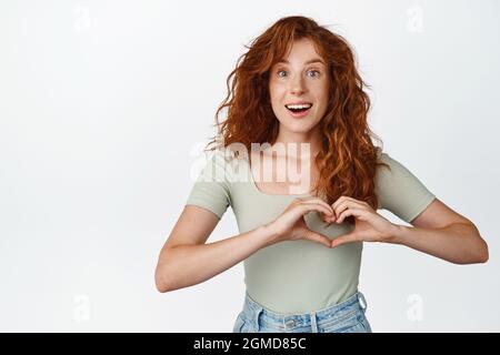 Jolie fille rouge montre le signe du coeur et sourit à l'appareil photo, se tient dans le t-shirt sur fond blanc Banque D'Images