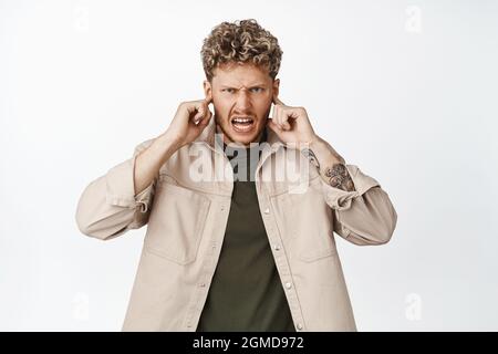 Un homme en colère et agacé ferme ses oreilles avec les doigts, regardant furieux à l'appareil photo, déteste le bruit fort, la musique terrible, se tenant sur fond blanc Banque D'Images