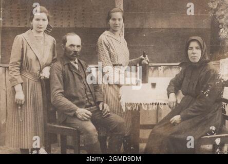 photo ancienne originale sur une famille de fermiers des années 1910. La famille assis autour d'une table sur la terrasse et tandis que l'une des filles tient une bouteille de boissons pour verser de l'alcool, de la palinka, du whisky, du roum... à ses parents jusqu'à ce que l'autre fille tient un rosaire pour prier DIEU! Banque D'Images