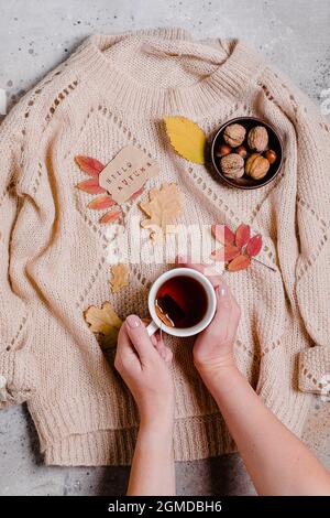 Tasse de thé chaud dans les mains de la femme. Fond d'automne en béton avec feuilles et confortable pull beige. Banque D'Images