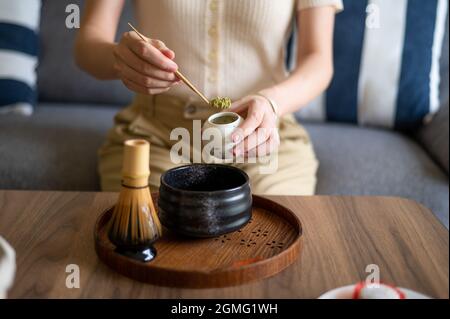 Femme qui fait une boisson japonaise au thé vert matcha à la maison en utilisant un set traditionnel de cérémonie du thé en bambou tout en étant assise sur le canapé à la maison. Banque D'Images