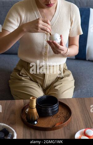 Femme qui fait une boisson japonaise au thé vert matcha à la maison en utilisant un set traditionnel de cérémonie du thé en bambou tout en étant assise sur le canapé à la maison. Banque D'Images