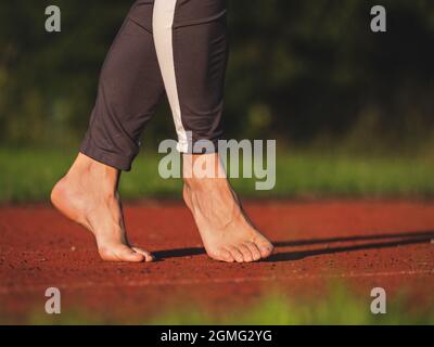 La femme adulte pieds nus marche sur la pointe du pied, étirant les tendons et les chevilles sur la piste de course rouge au soleil du soir. Corps d'âge moyen avec longs leggings Banque D'Images