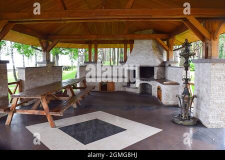 Grand belvédère dans le parc avec un toit en bois et un sol en mosaïque de pierre. A l'intérieur il y a une table en bois, un four en pierre et un cintre pour les vêtements. Banque D'Images