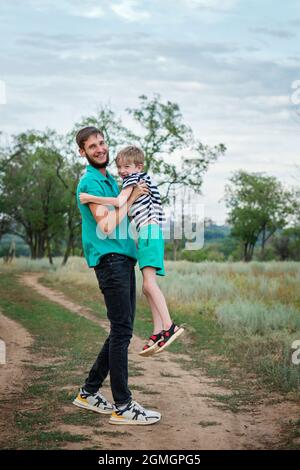 Portrait de famille authentique sur toute la longueur. Jeune papa décontracté avec son fils qui se serre et regarde dans l'appareil photo. Père et enfant marchant dans la campagne. La vie quotidienne. Activités quotidiennes. Banque D'Images