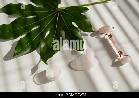 Grattoir de gouache rose, rouleau, pot de crème de feuille verte sur une table blanche. Équipement pour l'auto-massage et les soins de la peau pour le visage et le cou Banque D'Images