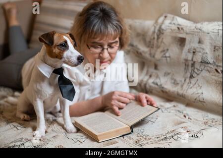 Une femme caucasienne âgée est allongée sur un canapé avec un chien intelligent Jack russell terrier portant des lunettes et une cravate et lisant un livre. Banque D'Images