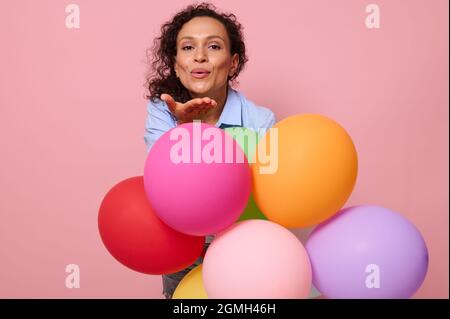 Charmante jeune femme afro-américaine envoyant un baiser aérien à la caméra, debout derrière des ballons multicolores sur fond rose avec espace de copie. Être Banque D'Images