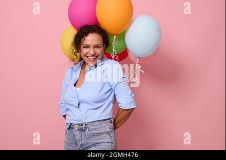 Charmante femme africaine joyeuse en chemise bleue riant et souriant avec un sourire crasseux regardant la caméra, tenant des ballons d'air multicolores derrière lui Banque D'Images