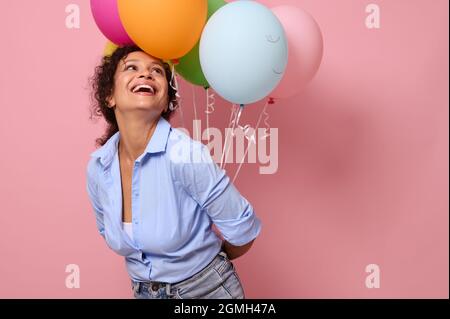 Joyeuse et joyeuse jeune femme africaine en chemise bleue riant et souriant avec un sourire crasseux regardant des ballons d'air multicolores dans ses mains Banque D'Images