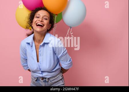 Joyeuse jeune femme latino-américaine avec des émotions joyeuses et heureuses riant en regardant la caméra, tenant des ballons derrière elle, isolée sur colo rose Banque D'Images