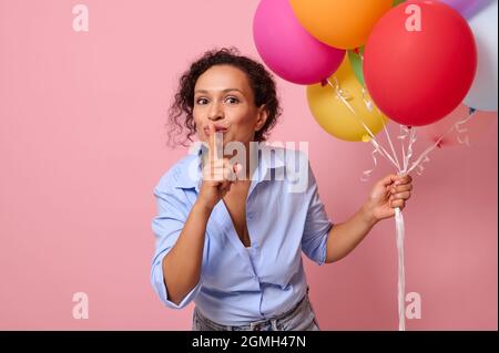 Mystérieuse mignonne jeune femme mixte de race avec des ballons d'air derrière son dos met l'index sur ses lèvres , gestant CHUT. Concept de surprise isolé Banque D'Images