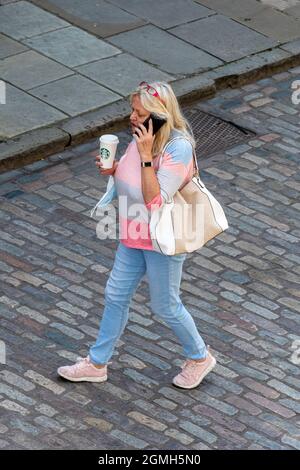 Femme marchant dans une rue pavée en haut parlant sur un téléphone portable, tenant une tasse de café Starbucks avec un masque de visage accroché à son poignet, 2021 UK Banque D'Images