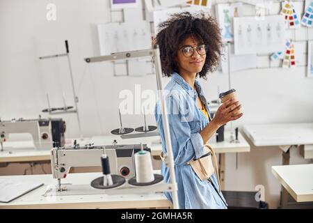 Une couturière afro-américaine avec une tasse et un sac à ceinture se penche sur une table avec une machine à coudre Banque D'Images