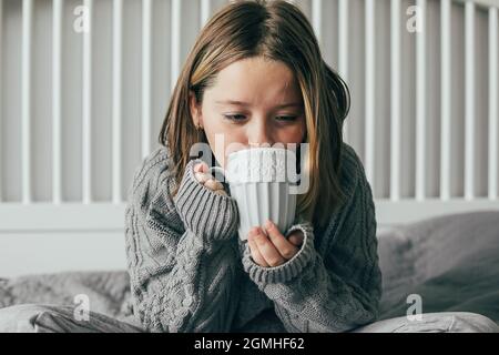 Une jeune fille boit du chocolat chaud sur un lit confortable dans la chambre. Concept de week-end d'automne ou d'hiver Banque D'Images