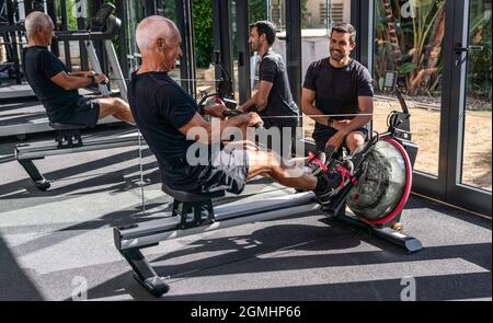 Un homme européen âgé, dans une salle de gym, avec un entraîneur personnel, en simulant l'aviron, sur un simulateur, à l'intérieur. Banque D'Images