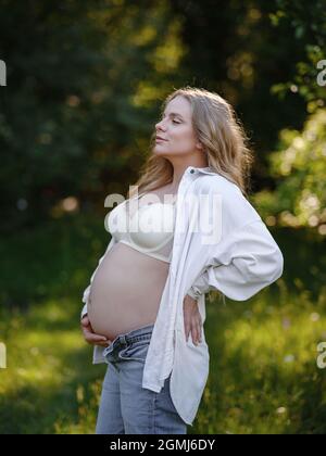 Femme blonde enceinte en chemise blanche et jeans apprécie le silence et la beauté de la nature. Idée de la santé et de la beauté de la grossesse Banque D'Images