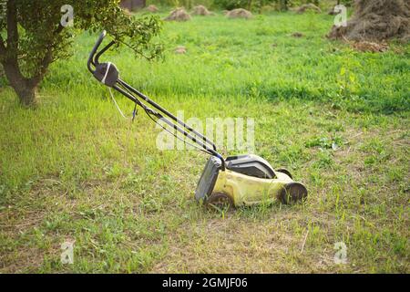 Le robot de tonte jaune sur roues repose sur une pelouse fauchée dans un jardin d'été. Poirier et tas avec de l'herbe sèche sur le fond Banque D'Images