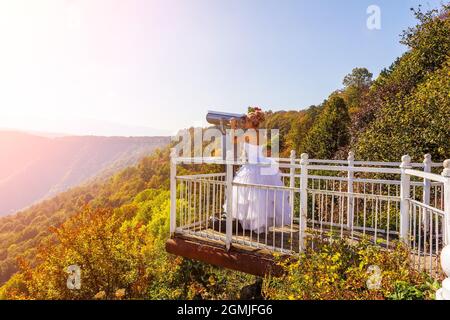 une mariée vêtir d'une robe blanche sur la terrasse d'observation regarde à travers des jumelles fixes binoscope au paysage de montagne le jour de l'automne Banque D'Images