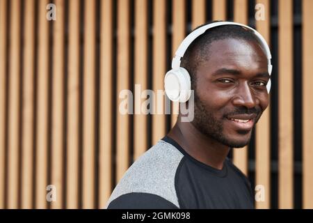 Portrait d'un jeune africain dans un casque sans fil souriant à l'appareil photo tout en marchant dans la ville Banque D'Images