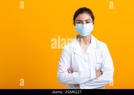 Portrait d'une femme médecin indienne portant une robe médicale blanche et un masque isolé sur fond jaune. Banque D'Images