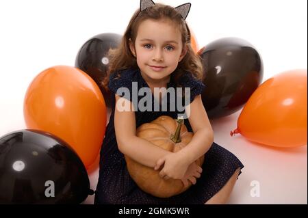 Portrait d'une charmante petite fille mignonne portant un cerceau avec des oreilles de chat, vêtu d'un costume de carnaval de sorcière foncé, chopes d'une citrouille dans sa main, asseyez-vous sur un whi Banque D'Images