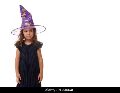 Portrait d'une jolie petite fille portant un chapeau de sorcier et vêtue d'une élégante robe de carnaval, regardant l'appareil-photo posant avec des bras croisés contre la ba blanche Banque D'Images