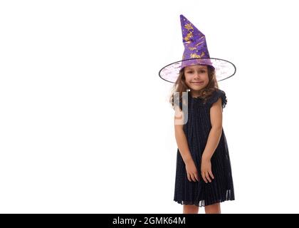 Portrait d'une jolie petite fille portant un chapeau de sorcier et vêtue d'une élégante robe de carnaval, regardant l'appareil-photo posant avec des bras croisés contre la ba blanche Banque D'Images