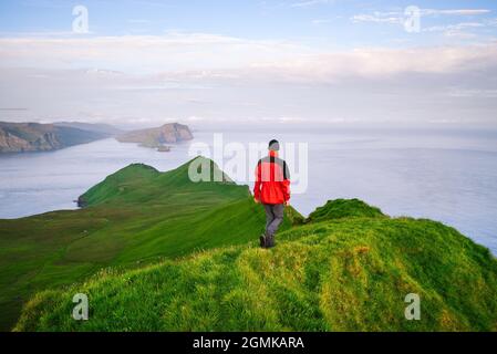 Un touriste dans une veste rouge marche à travers les collines vertes sur l'île de Mykines, îles Féroé Banque D'Images