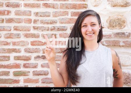 Femme souriante avec de longs cheveux faisant un signe de victoire avec sa main sur un mur de brique. Copier l'espace pour le texte Banque D'Images
