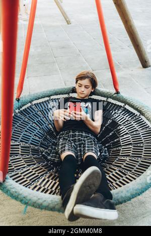 Adolescente utilisant le téléphone portable sur l'oscillation des enfants dans le parc Banque D'Images