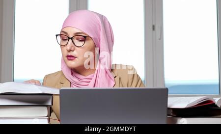 Femme dans hijab et lunettes dactylographiant sur ordinateur portable regardant dans des livres Banque D'Images