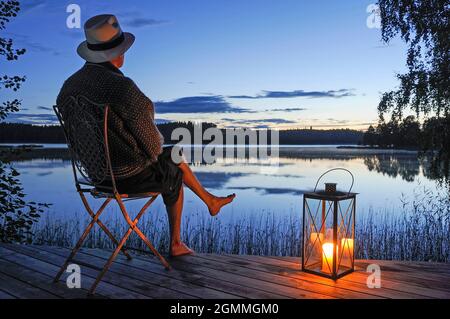 Un homme assis sur une terrasse en bois au bord d'un lac le soir Banque D'Images