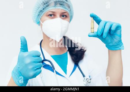 Un médecin dans un masque médical et des gants tient une ampoule avec un vaccin dans sa main et montre un semblable. Bravo. Un antidote fiable et éprouvé à la maladie. Banque D'Images