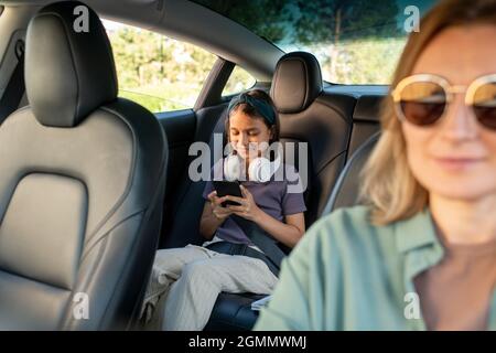 Bonne écolière avec un casque défilant dans un téléphone intelligent tout en étant assise sur le siège arrière de la voiture Banque D'Images