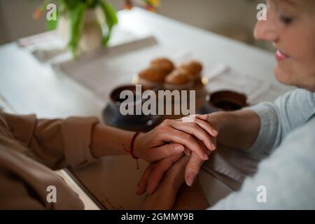 Gros plan de la mère âgée tenant les mains avec la fille adulte à l'intérieur à la maison, prenant du café. Banque D'Images