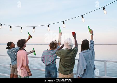 Vue arrière des jeunes amis avec des boissons devant le bord de l'eau le jour de l'été Banque D'Images