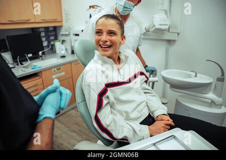 Cliente caucasienne souriante assise sur le lit du dentiste discutant avec les médecins après une intervention chirurgicale Banque D'Images