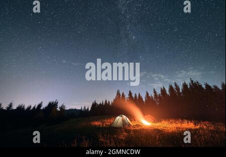 Campez de nuit sous un ciel étoilé. Vue grand angle sur un paysage magnifique en montagne. Deux tentes touristiques et feu de camp, mur de spruces en arrière-plan. Concept de voyage et d'astrophotographie Banque D'Images