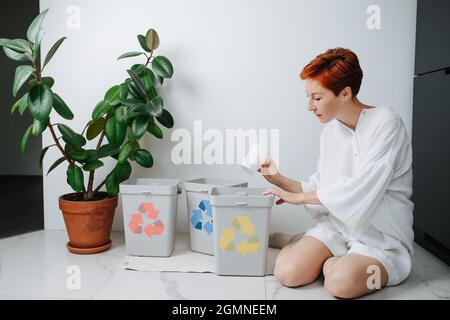 Femme préoccupée assise sur ses jambes, triant les ordures dans de petites poubelles de recyclage à la maison. Ils ont des flèches de couleur différente. Plastique de maintien Banque D'Images