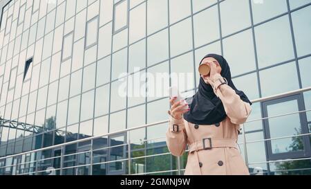 Jeune femme musulmane dans hijab tenant un smartphone et buvant du café pour aller près du bâtiment Banque D'Images