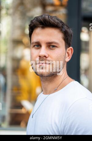 Beau jeune homme avec une coupe de cheveux élégante et des poils portant t-shirt blanc regardant l'appareil photo tout en se tenant contre un arrière-plan urbain flou Banque D'Images